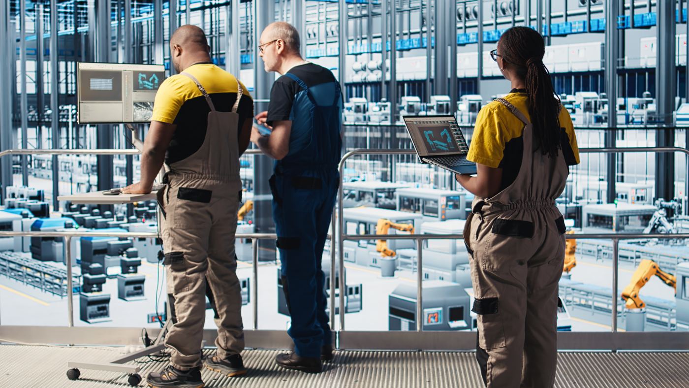 Drie technici overzien vanaf een bordes een productiehal, met een laptop en een beeldscherm